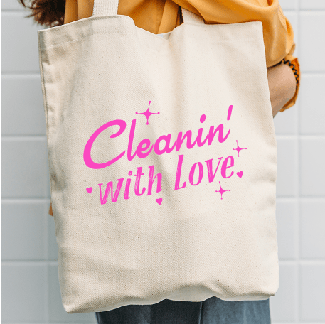 A person holding a beige canvas tote bag with pink text that reads 'Cleanin' with Love' surrounded by small heart and sparkle graphics.