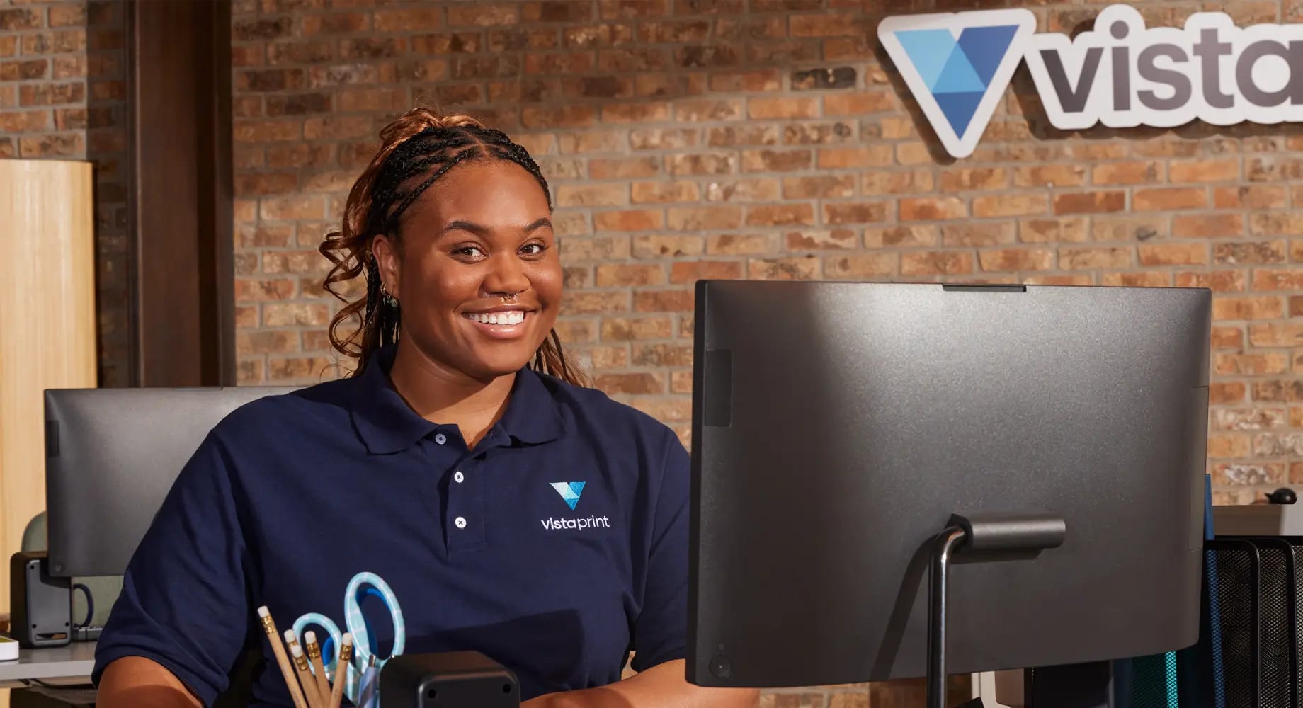 A friendly Vistaprint employee with braided hair smiles warmly while seated at a desk with a computer monitor, backed by a stylish brick wall and Vistaprint signage.