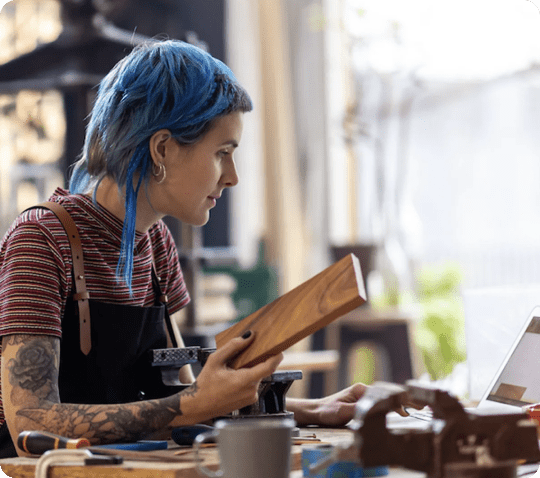 Tattooed person with blue hair holding a piece of wood while working on a laptop in a workshop.