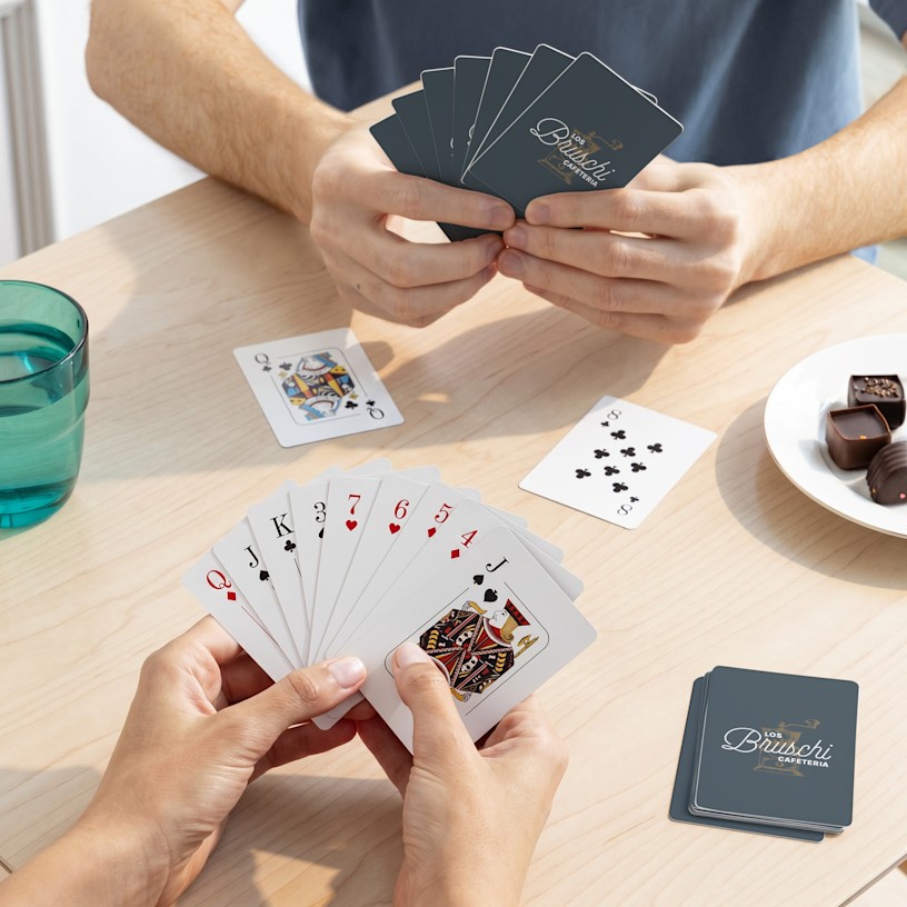 Two individuals playing cards at a wooden table laid out with snacks. 