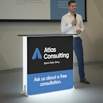 An illuminated lightbox display counter promoting a consultancy business, with a presenter standing beside it.