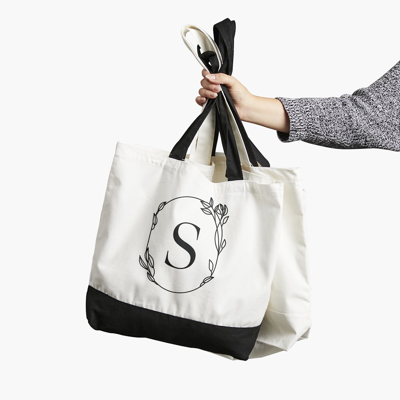 A person holding two custom tote bags with a floral design and a large 'S' in the center, featuring a black bottom and light-colored top.
