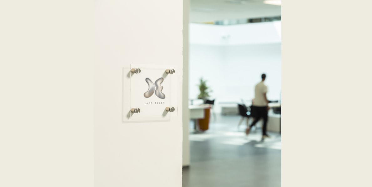 Ein quadratisches Firmenschild aus Acryl mit Logo an einer weißen Tür eines Büros.