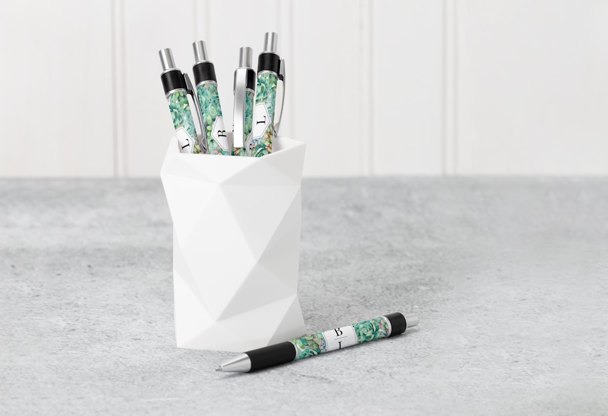 Four ballpoint pens with a floral design rest in a geometric pencil holder while a fifth ballpoint pen sits on a desk.