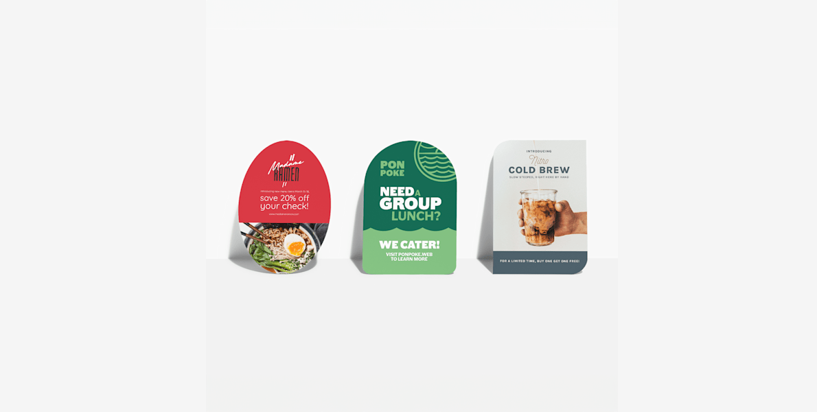Oval, half-circle and leaf-shaped postcards, all propped up against a wall and advertising different restaurants.