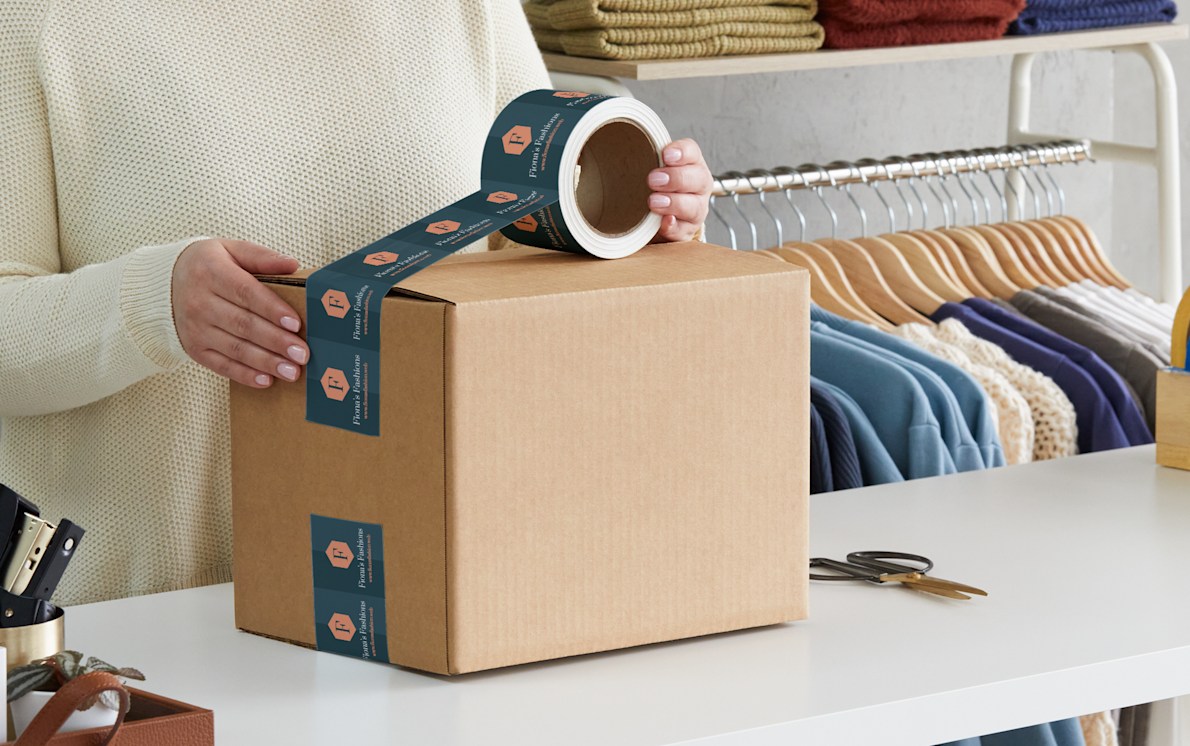 A woman in a white sweater applying white plastic Self-adhesive packaging tape to a cardboard box. The tape is custom printed with a design for a clothing and fashion business.