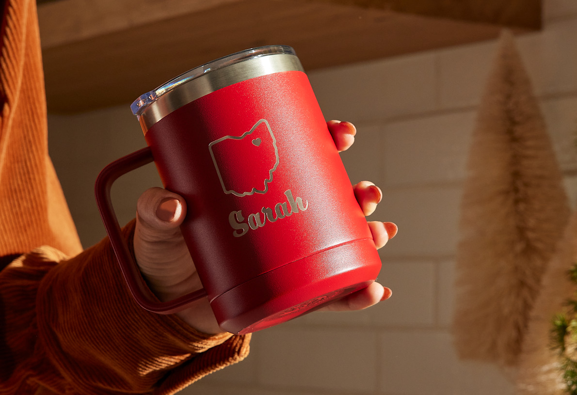 A hand holding a 15 oz. red travel mug at an angle. The mug is laser etched with the name Sarah and the outline of a US state. 