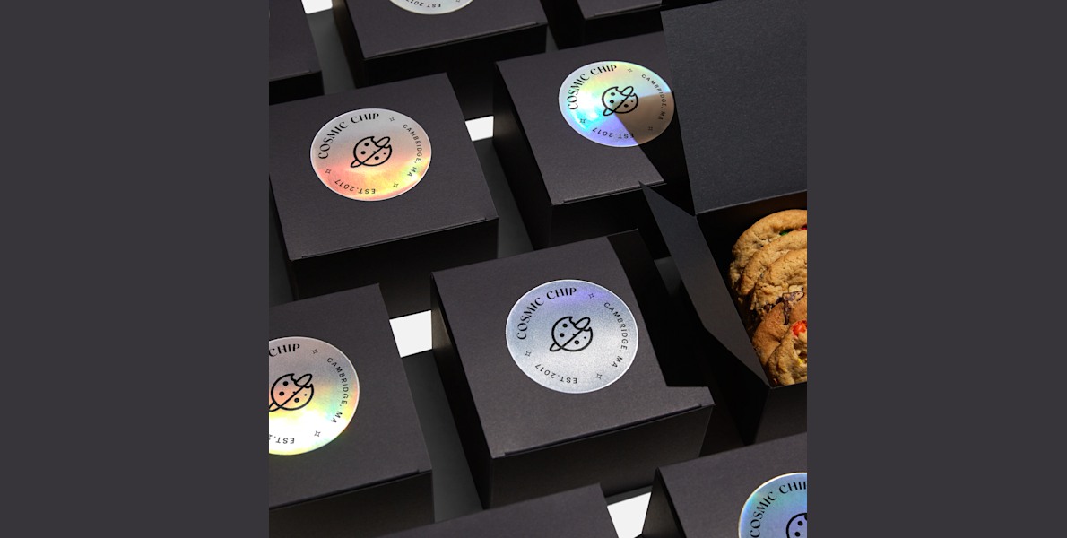 Black boxes neatly lined up in rows with circle holographic paper sticker labels applied to the top of each. One box is opened revealing cookies inside