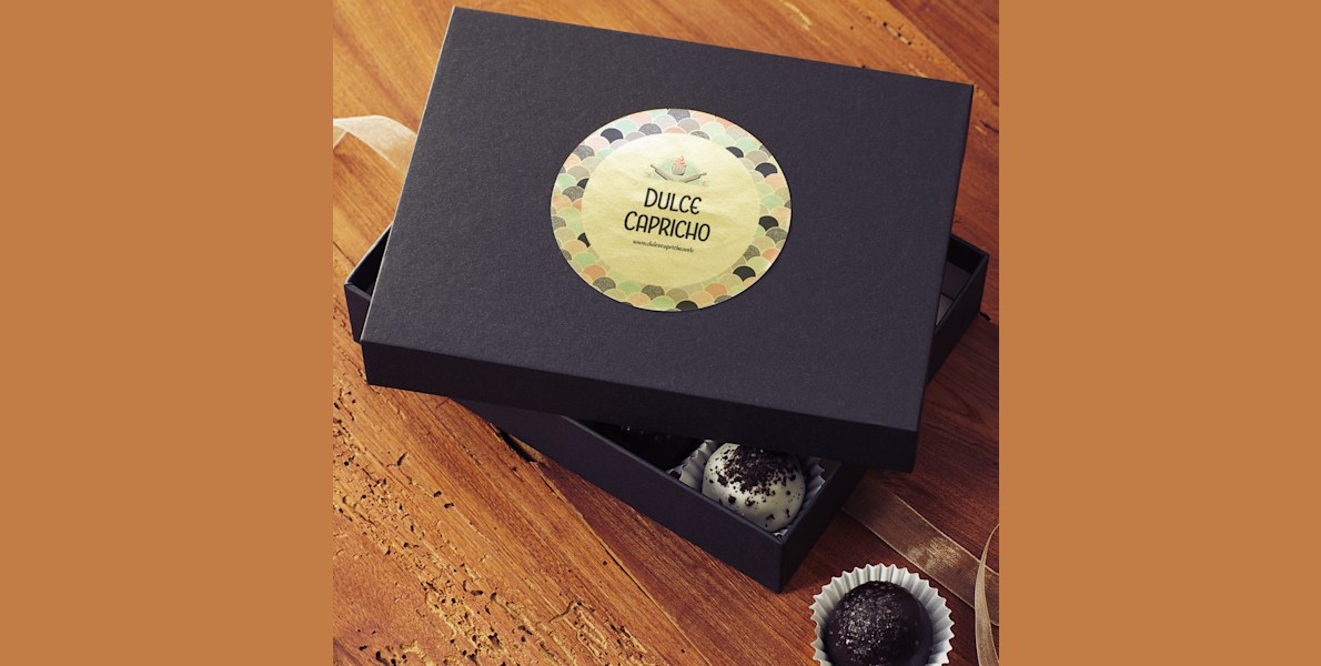 A black box sitting on a wooden table with the cover off revealing bon bons inside. The lid has a circular gold paper label applied to it.