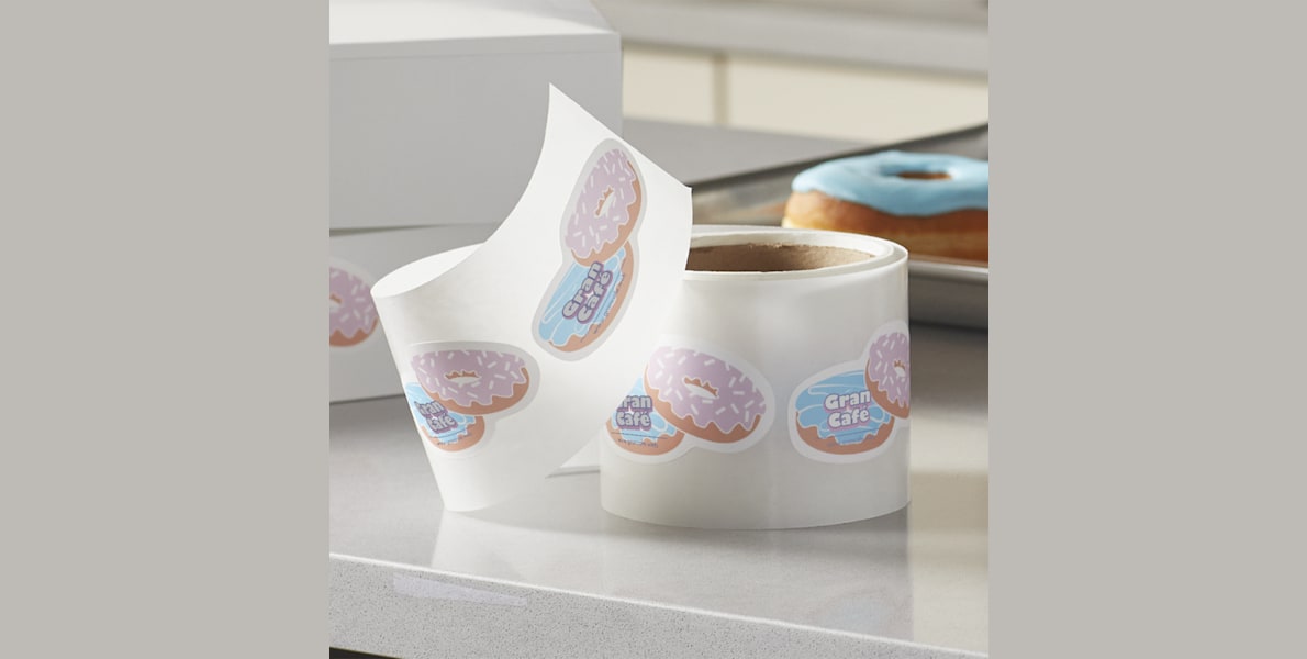 A roll of die-cut label stickers in the shape of two doughnuts in an offset stack placed on a countertop infront of two boughnut boxes, and a tray of doughnuts.