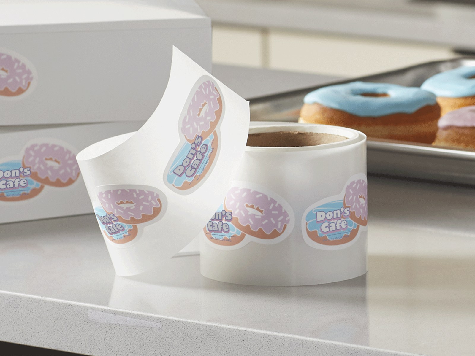 A roll of die-cut label stickers in the shape of two doughnuts in an offset stack placed on a countertop infront of two boughnut boxes, and a tray of doughnuts.