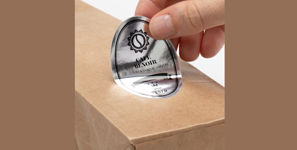 A bag of coffee on a gray background with a hand applying a silver paper oval label to the front.