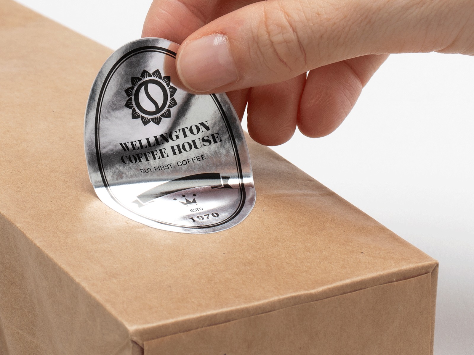 A bag of coffee on a gray background with a hand applying a silver paper oval label to the front.