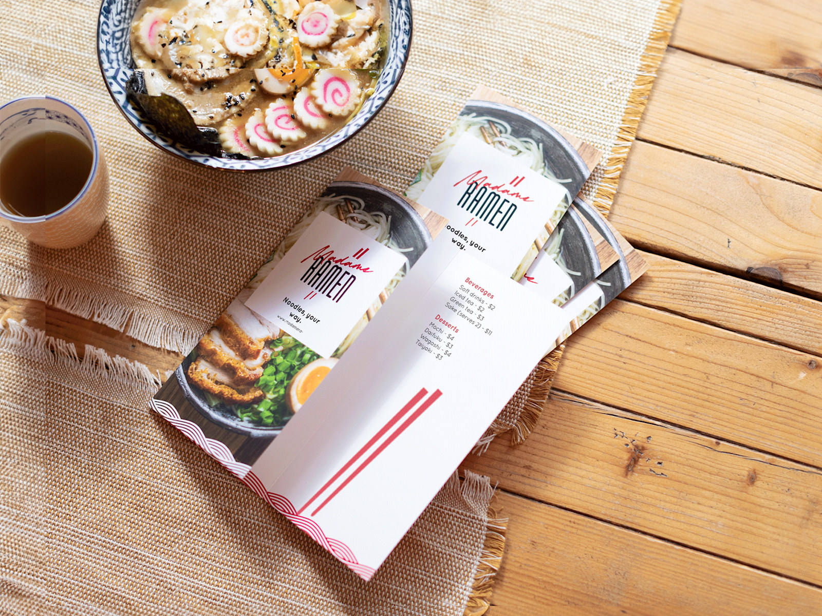 A small stack of tri-fold menus for a ramen restaurant, sitting next to a cup of tea and bowl of soup. 