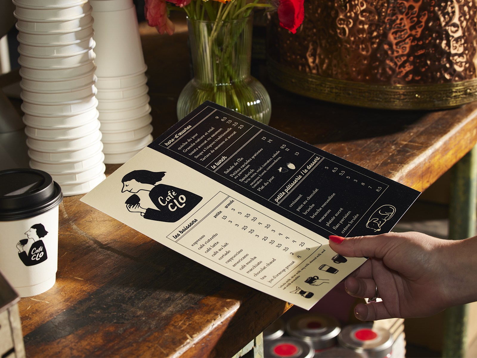 A flat menu for a coffee shop, held by a customer in front of a stack of custom cups. 