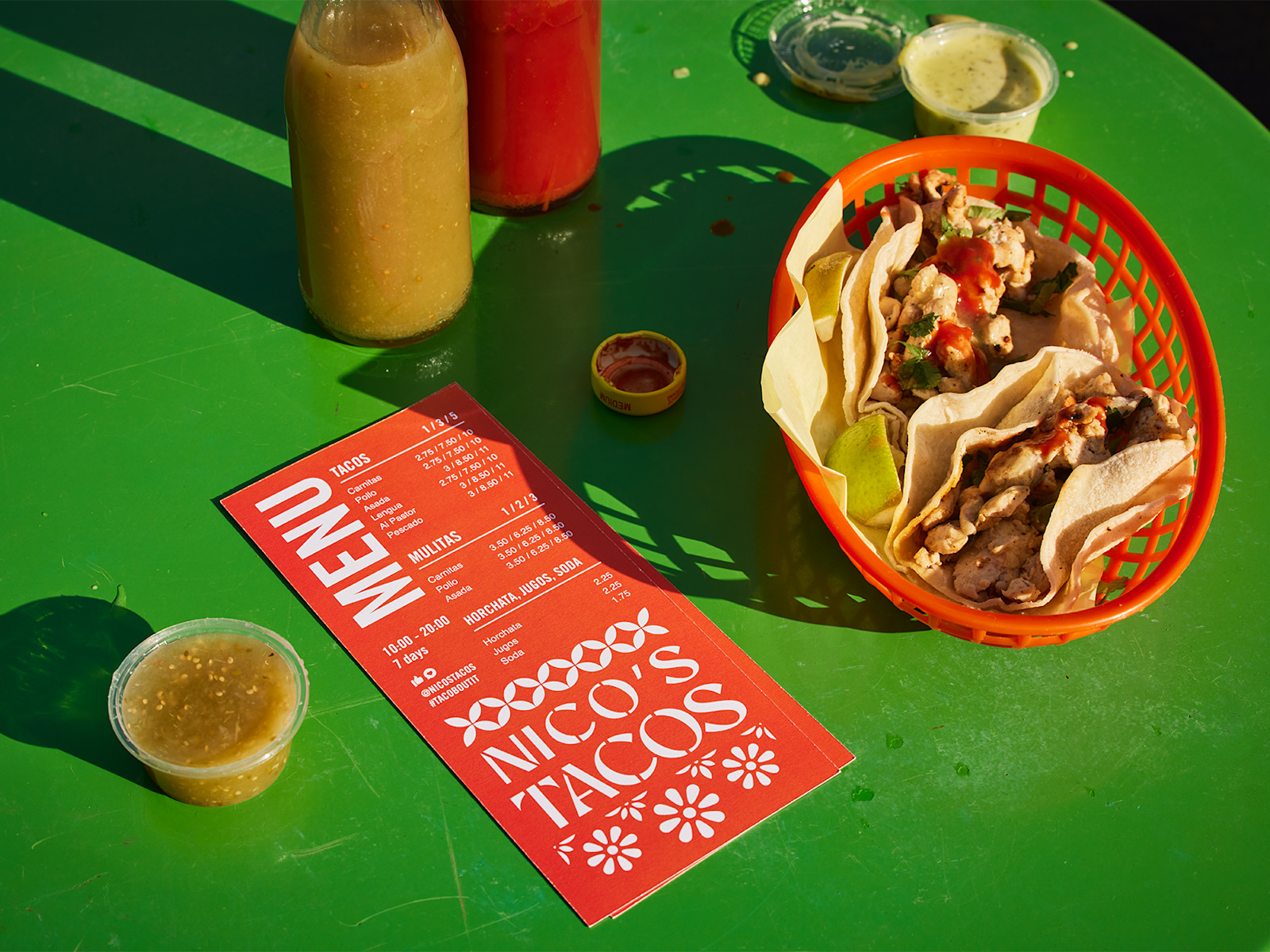 A flat menu for a taco restaurant, on a table next to a basket of tacos and sauce containers.