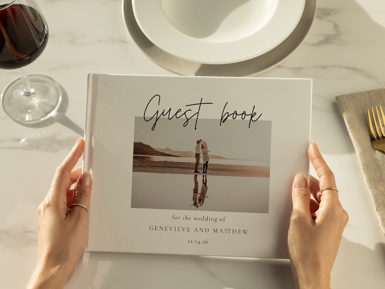 Larger version: A pair of hands holding a wedding guest book, with an elegant tablesetting in the background. 