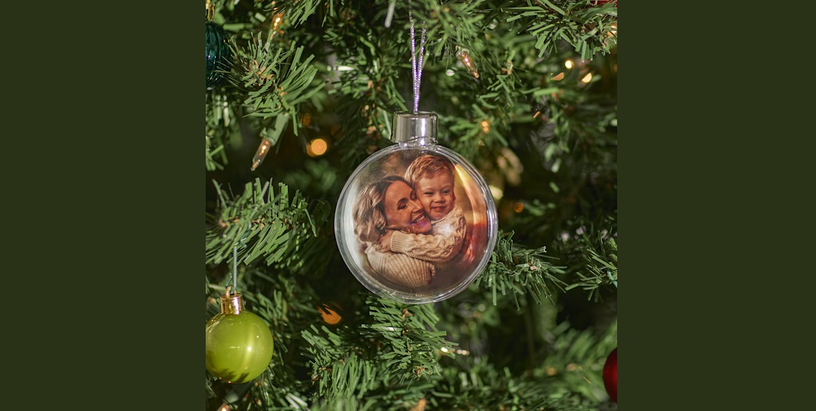 Aan een kerstboom hangt een kerstbal met foto, gepersonaliseerd met een foto van een moeder en haar zoon die elkaar omhelzen. 