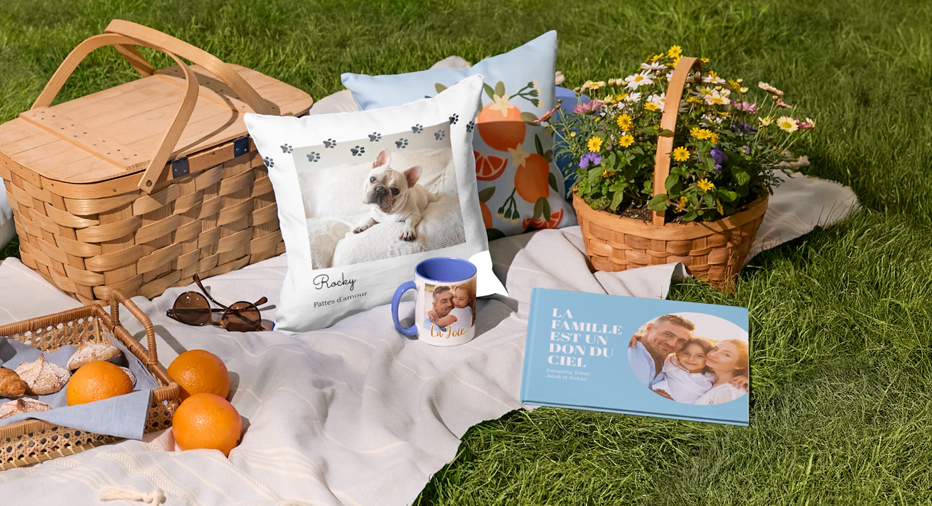 Une tasse, un oreiller et un livre photo personnalisés disposés sur une couverture près d’un panier de pique-nique.