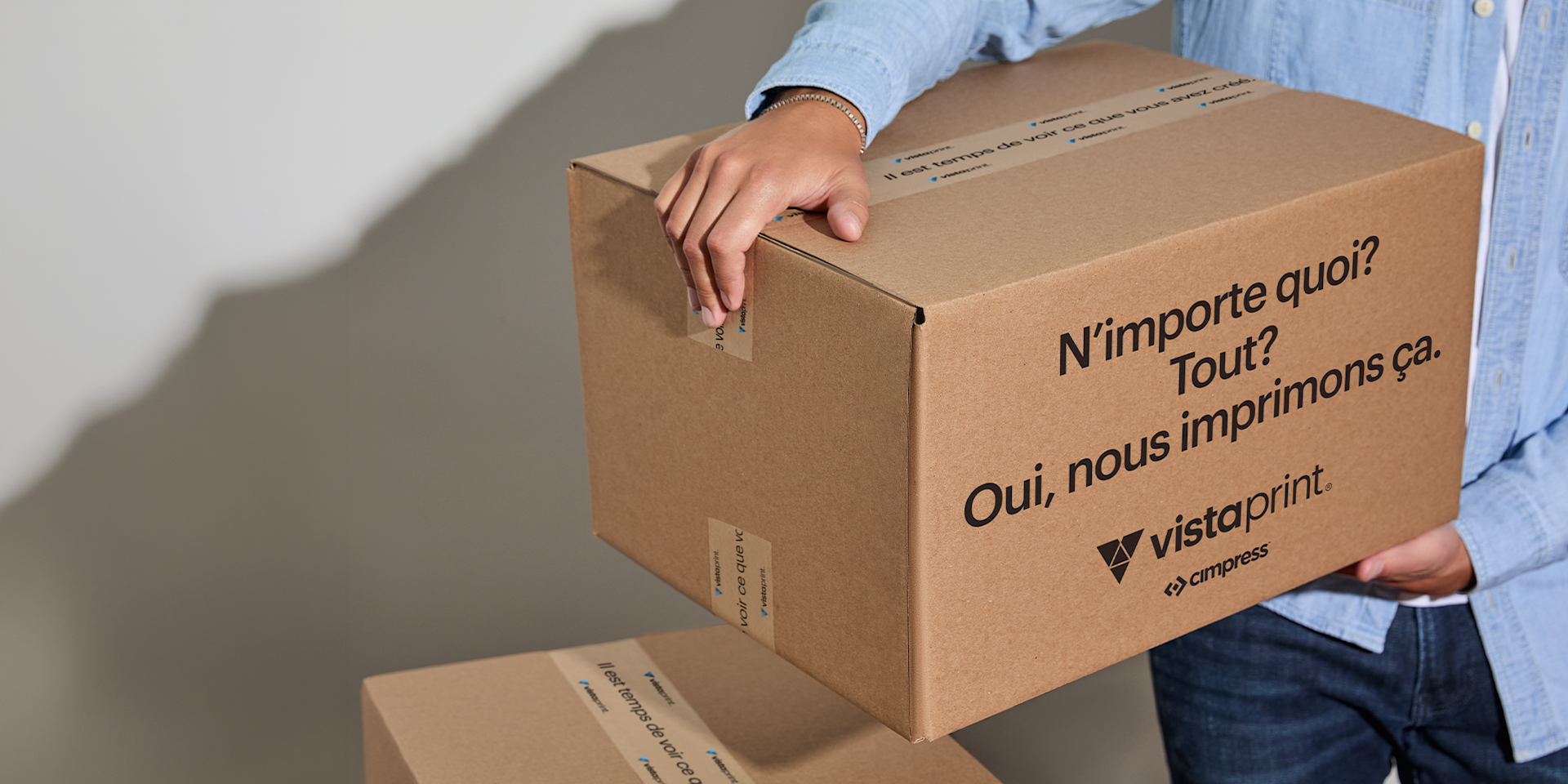 Personne tenant une boîte d’expédition en carton aux couleurs de VistaPrint, illustrant la rapidité des délais de production.