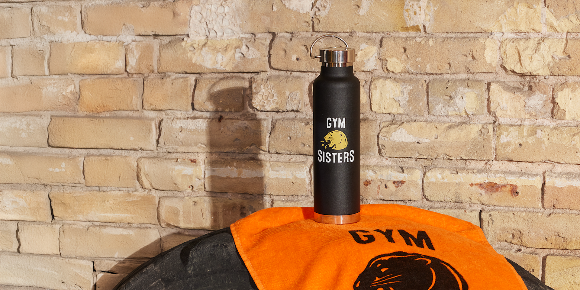 A black reusable water bottle labeled “Gym Sisters” placed on an orange gym towel against a brick wall background. 