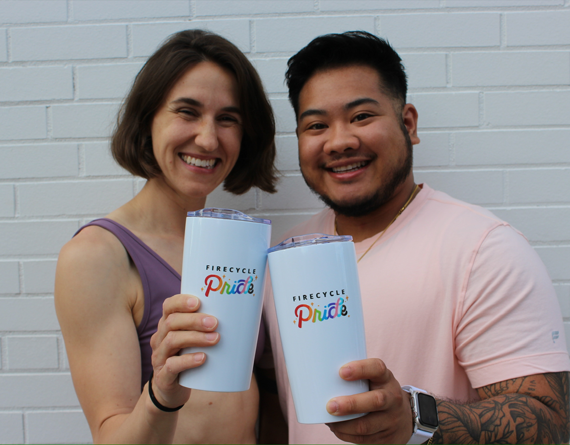 Two happy FireCycle members stand side by side against a plain white brick wall, smiling warmly while holding up their personalized FireCycle Pride tumblers. Their relaxed and cheerful expressions perfectly capture the spirit of unity and pride.
