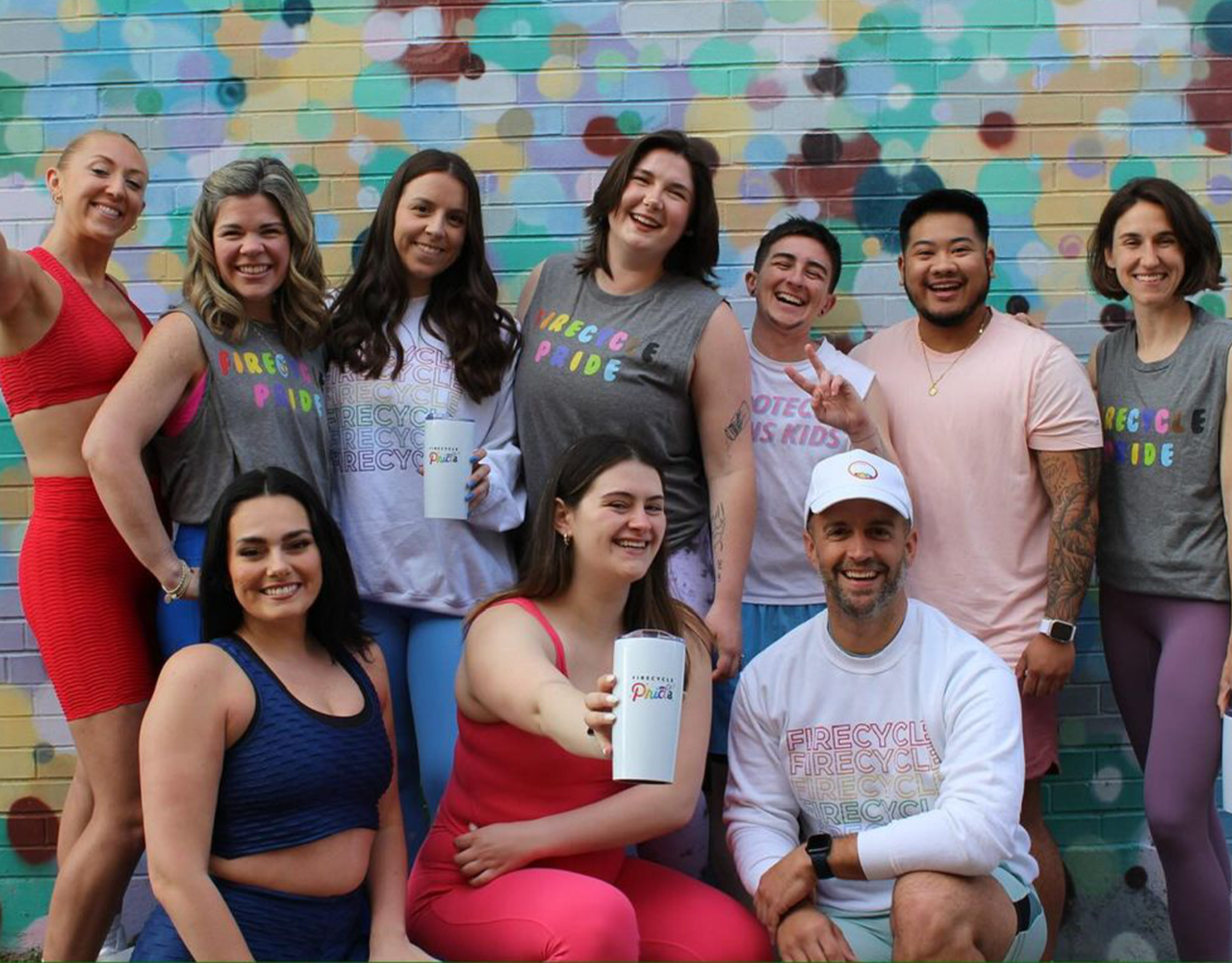 A diverse and joyful group of FireCycle members gather in front of a colorful mural, all smiling and proudly holding white tumblers personalized with the FireCycle Pride logo.