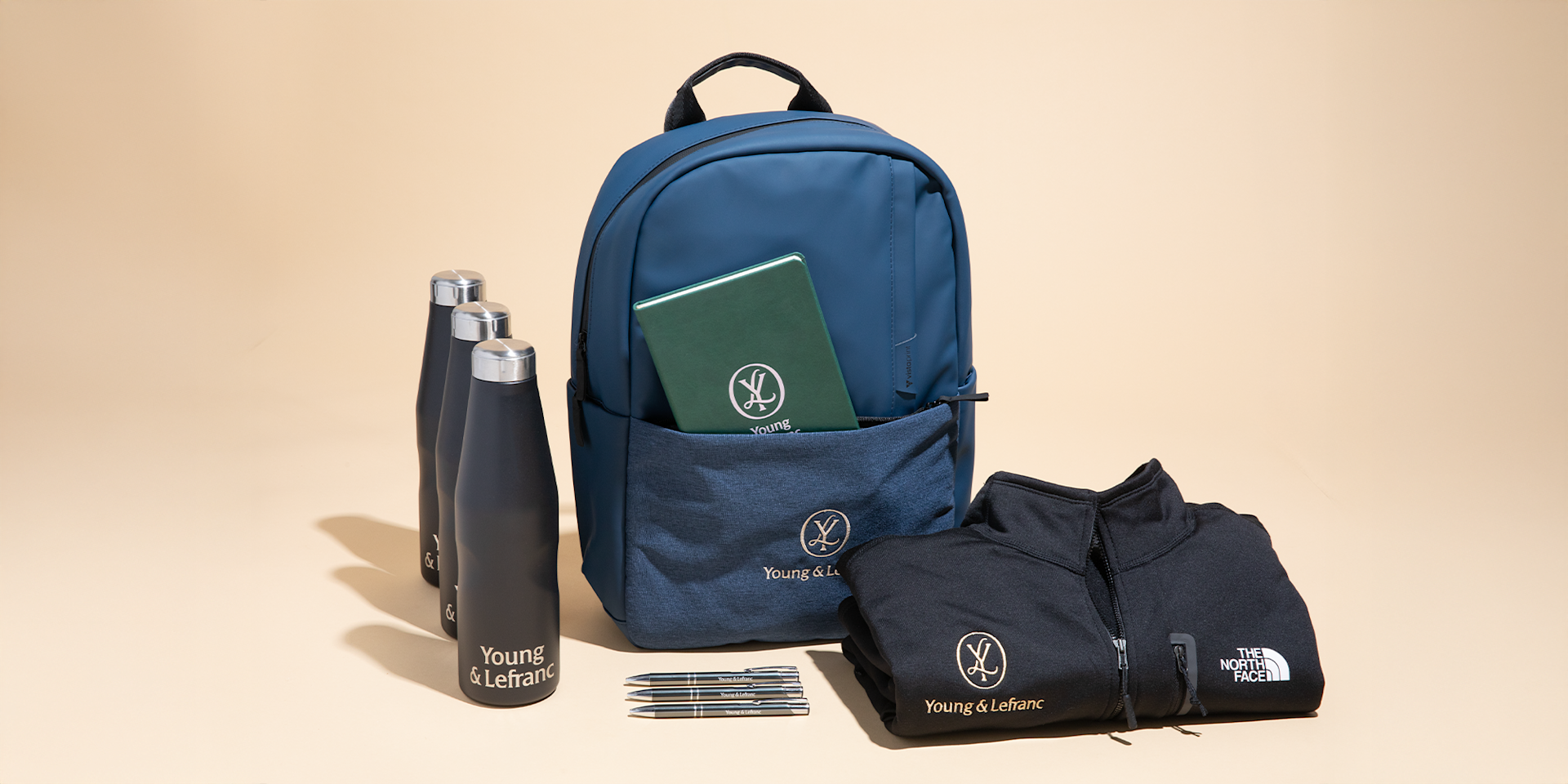 Mochila azul marino, botellas de agua negras, bolígrafos y ropa negra dispuestos juntos, todos con la marca personalizada Young & Lefranc.