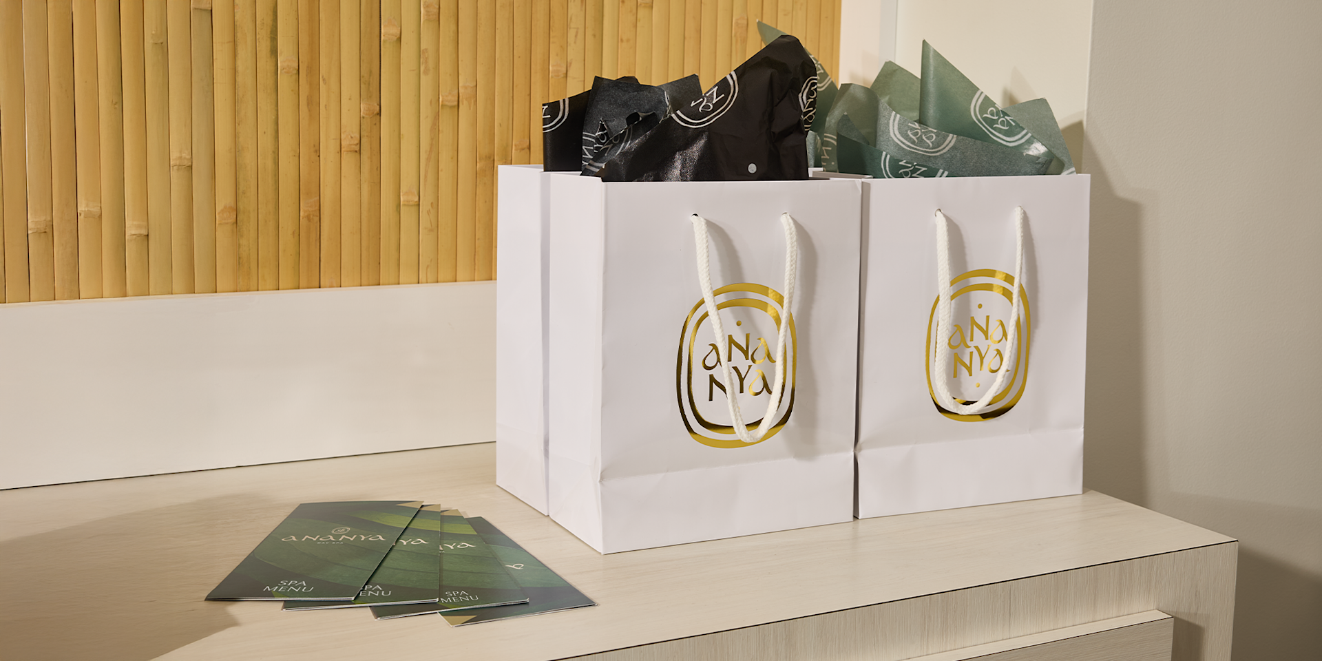 Primer plano de bolsas de papel blancas con logotipos dorados circulares, llenas de papel de seda verde salvia y negro que asoma por la parte superior.