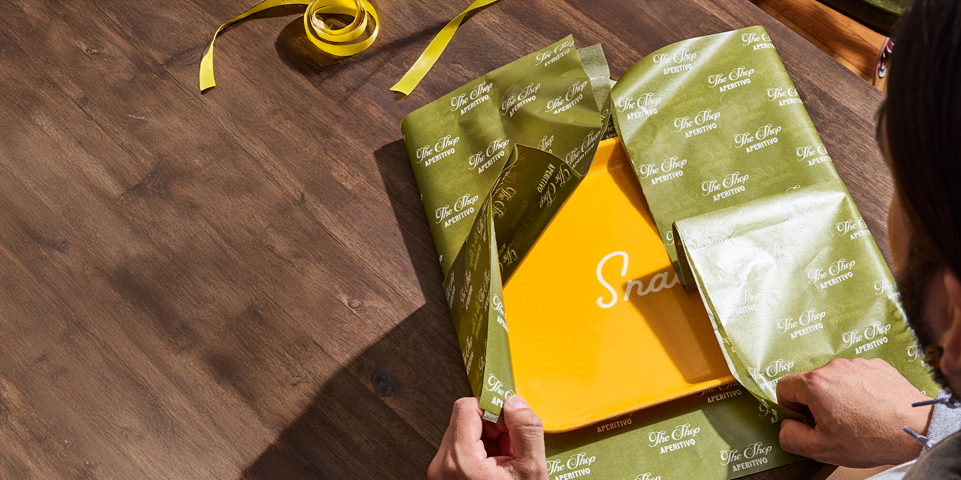 Mains emballant une boîte jaune dans un papier d’emballage vert de marque sur une table en bois, avec des ciseaux verts et un ruban jaune à côté.