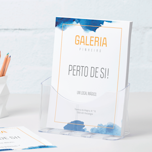 Um suporte de folhetos em plástico de tamanho A4 com flyers de uma galeria pousado numa secretária junto a um porta-lápis.