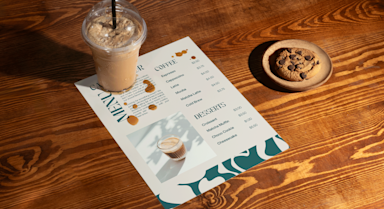 A flat synthetic menu on a cafe table with a frozen drink and cookie, undamaged by a small spill.