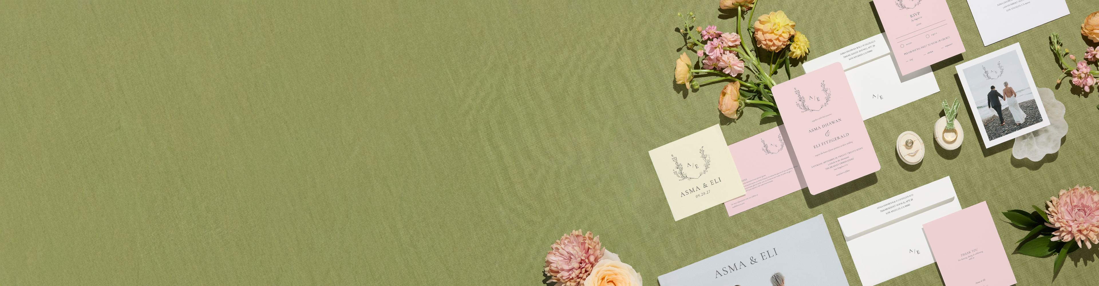 A wedding stationery suite elegantly arranged on a green tablecloth alongside some flowers and two engagement rings. 