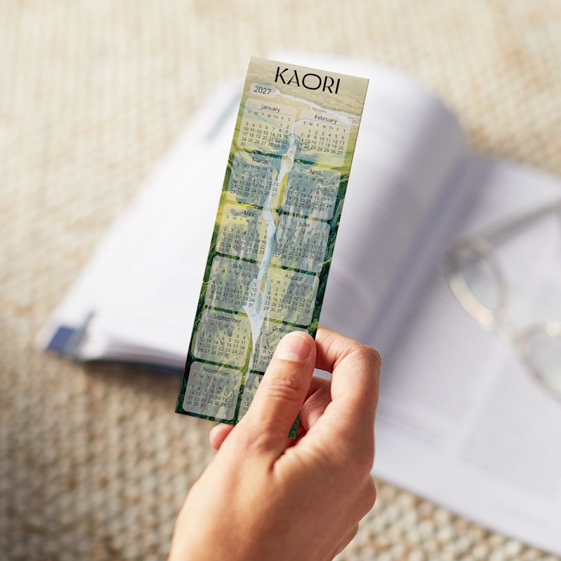 Someone holding a 2027 calendar bookmark with a green design, placed over an open book.