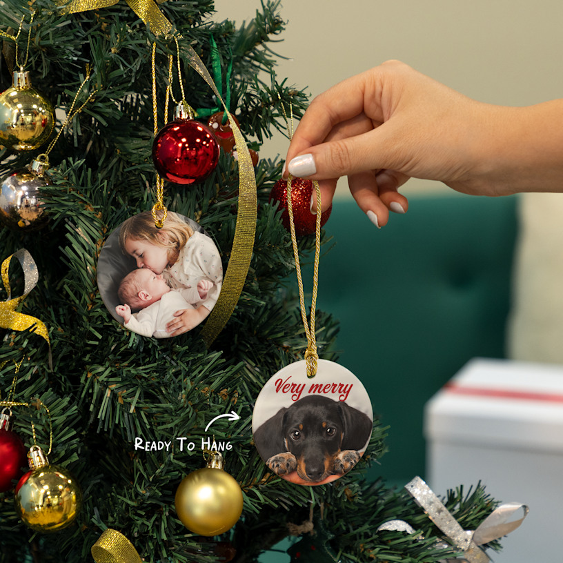 A Christmas ornament featuring a photo of two children, along with a festive greeting. An arrow points to the pre-drilled hole and gold string.