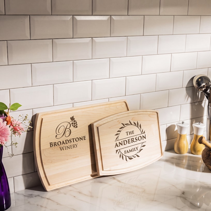 Two custom cutting boards leaning against a kitchen backsplash, one customized for a winery and the other with a family name. 