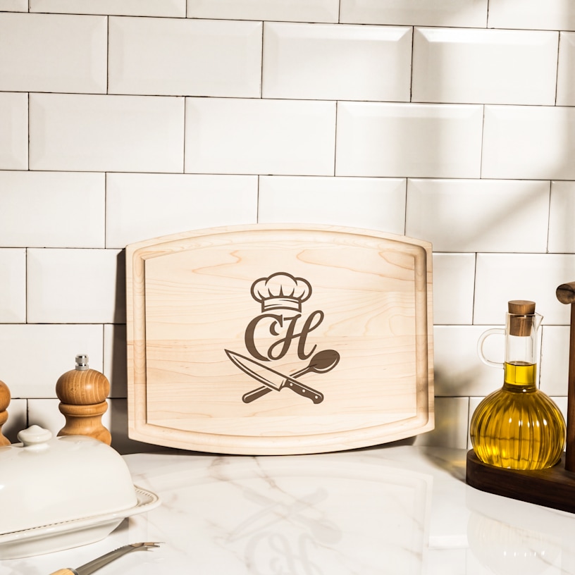 A custom cutting board etched with a monogram and chef graphics, leaning against a kitchen backsplash.