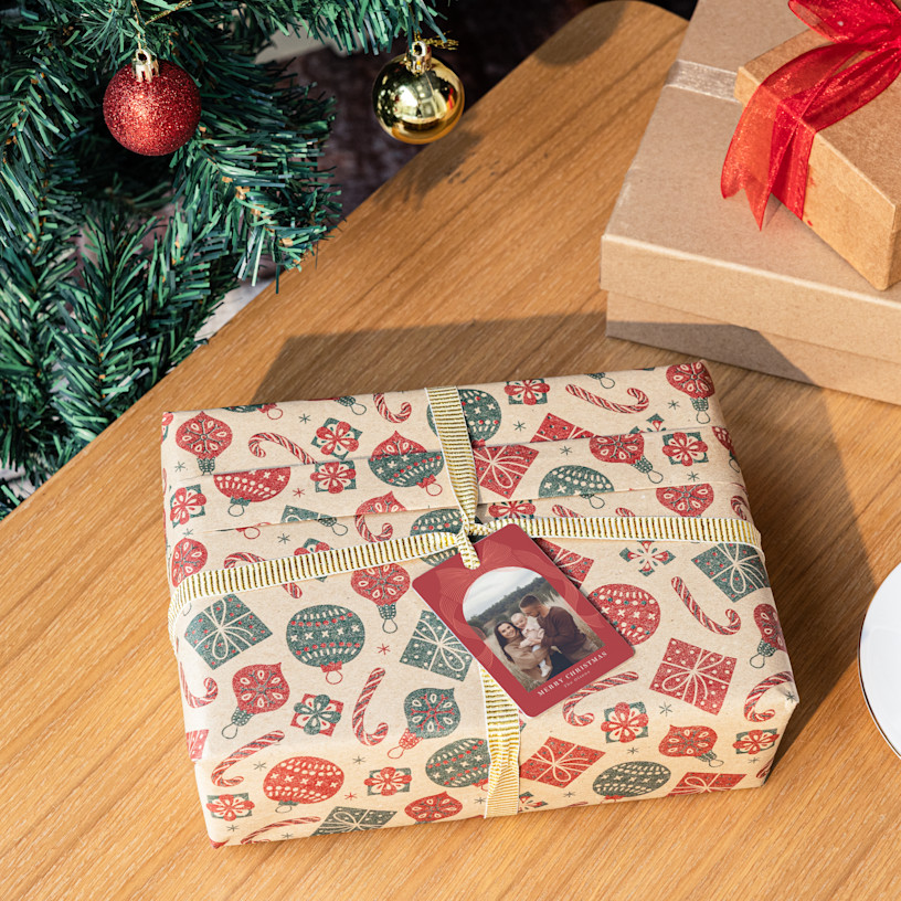 A wrapped present topped with a custom ornament and placed alongside a Christmas tree.