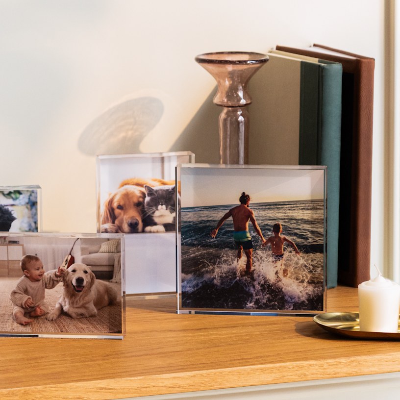 Tres bloques acrílicos de fotos en un estante de madera muestran fotos de mascotas, un niño con un perro y dos personas corriendo hacia el océano al atardecer.