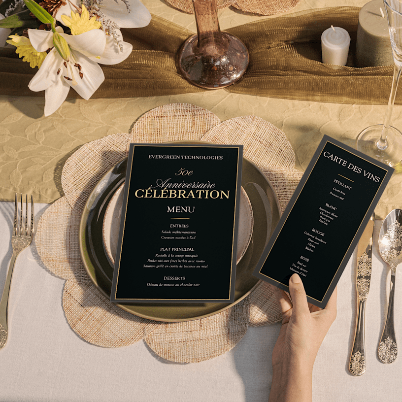 Deux élégants menus noirs et dorés pour un 50e anniversaire, présentés sur une table dressée avec soin, l’un posé sur une assiette et l’autre tenu par un invité. 