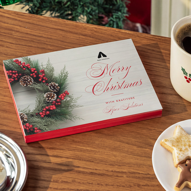 A stack of painted edge holiday invitations on a kitchen table next to a mug of hot cocoa. 
