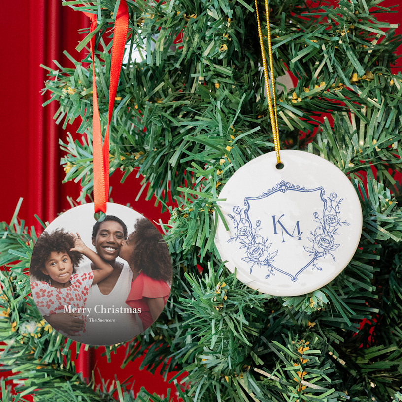 Two custom ornaments hanging from a Christmas tree, one printed with a monogram and the other with a photo of a mother and her two daughters.