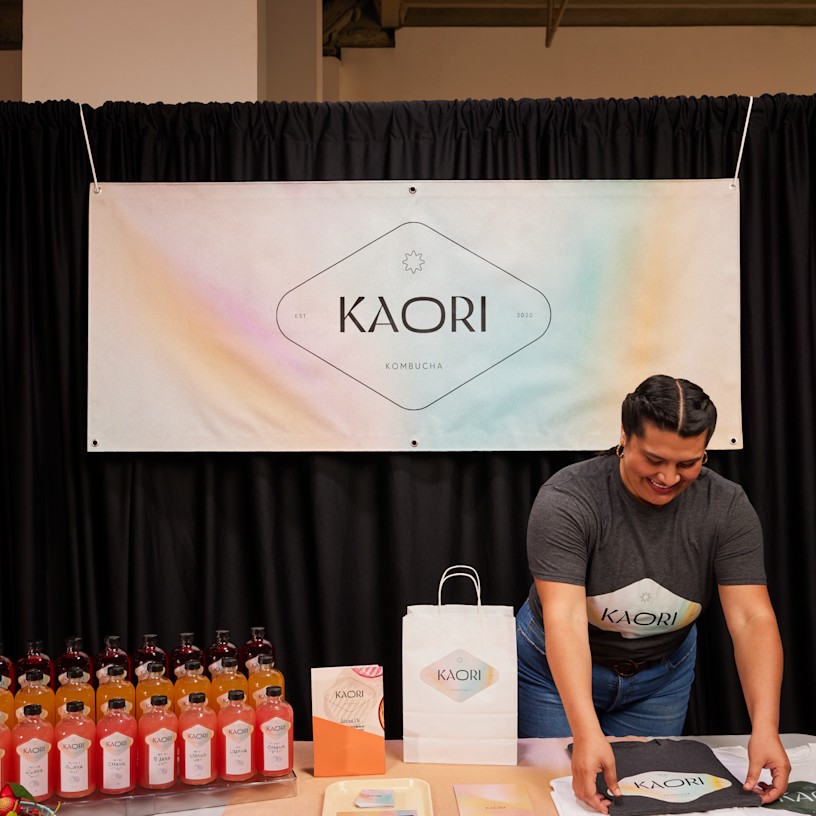 Et kombucha-brands salgsstand viser et flot standardbanner, drikkevarer, poser og reklamematerialer.