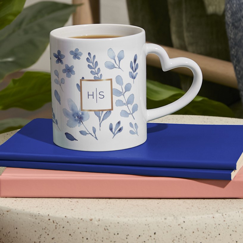 Et krus med en hjerteformet hank og et personligt monogram, der hviler på en stak bøger.  