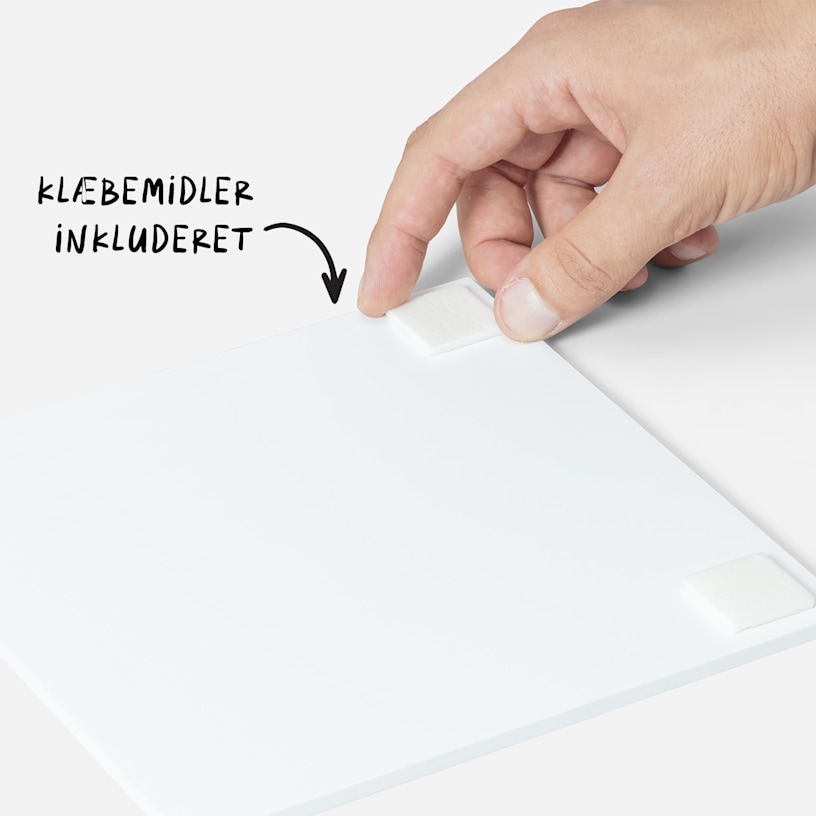 En hånd, der placerer en dobbeltsidet klæbestrimmel på bagsiden af en fotoflise, med en bemærkning om, at der følger klæbemiddel med fliserne. 