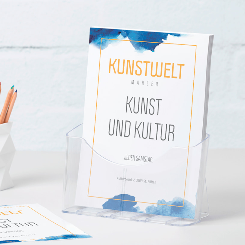 Ein DIN-A4-Flyerhalter aus Kunststoff mit Flyern einer Galerie auf einem Schreibtisch neben einem Stiftbecher.
