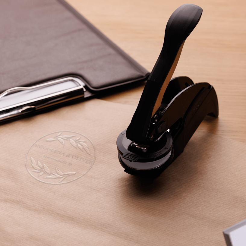 Ein Prägestempel neben personalisierten Schreibwaren aus Kraftpapier samt geprägtem runden Motiv.