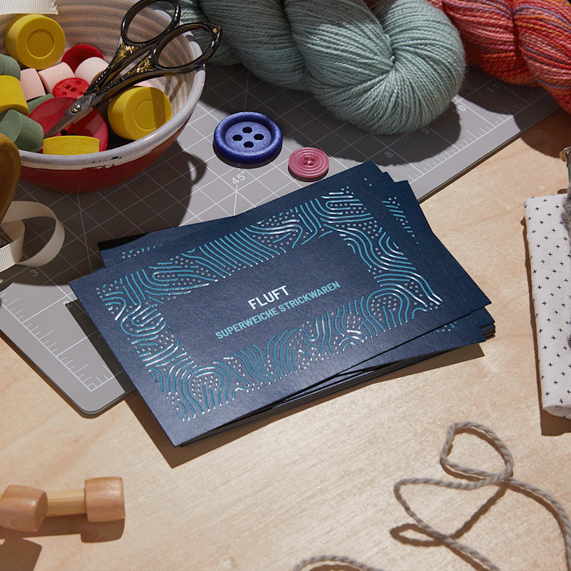 Eine Visitenkarte mit Spotlack für ein Strickwarengeschäft, umgeben von Strickzubehör.