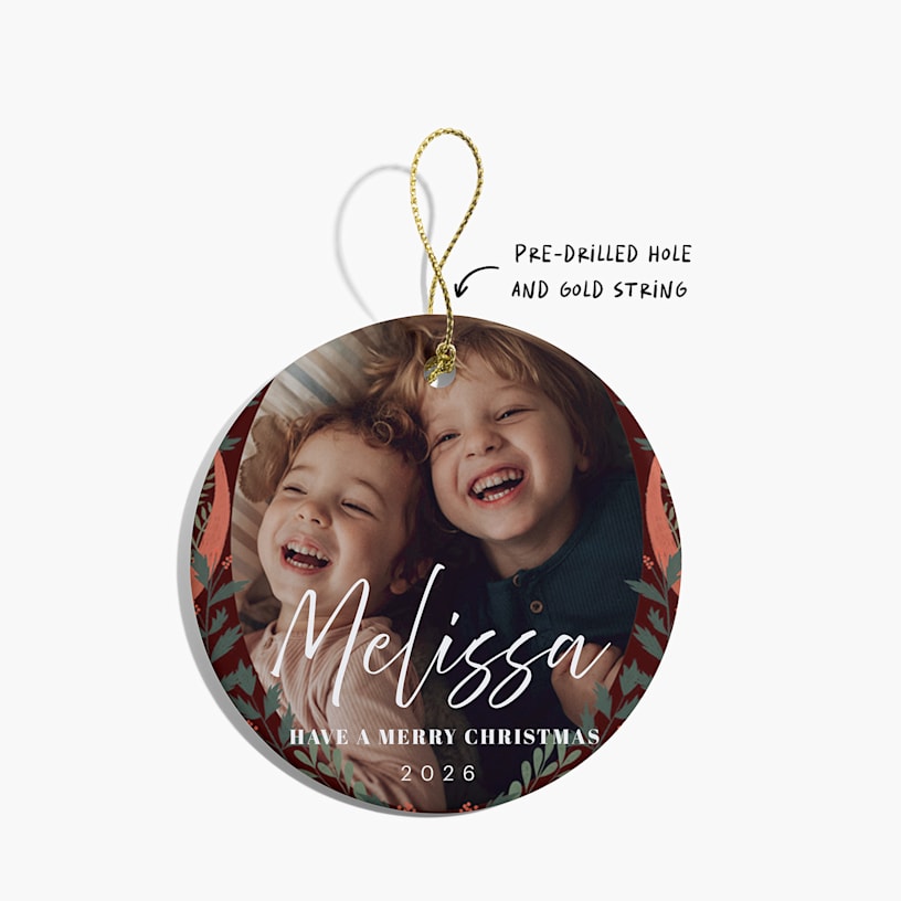 A Christmas ornament featuring a photo of two children, along with a festive greeting. An arrow points to the pre-drilled hole and gold string.