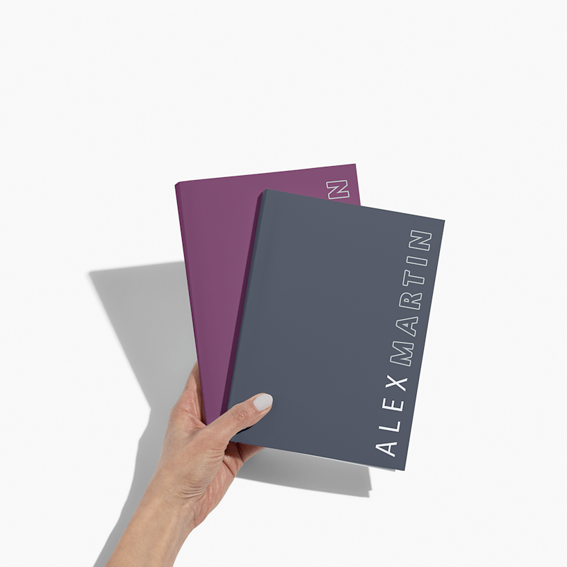 Two custom wraparound spiral notebooks in a person's hand, in maroon and navy. Both feature simple text on the right-hand edge of the cover.